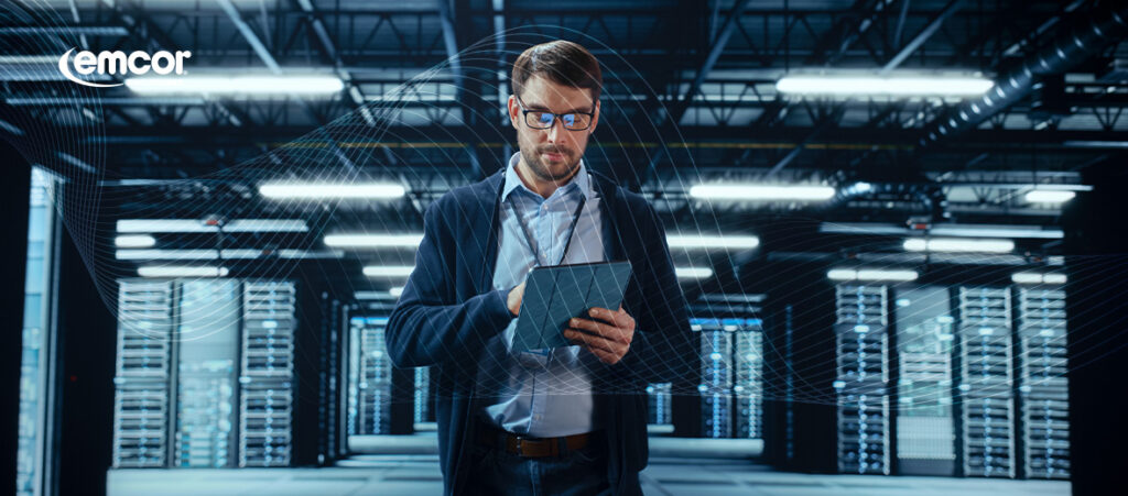 Professional engineer in business attire using a tablet in a modern data center facility with server racks visible in the background - Emcor branding